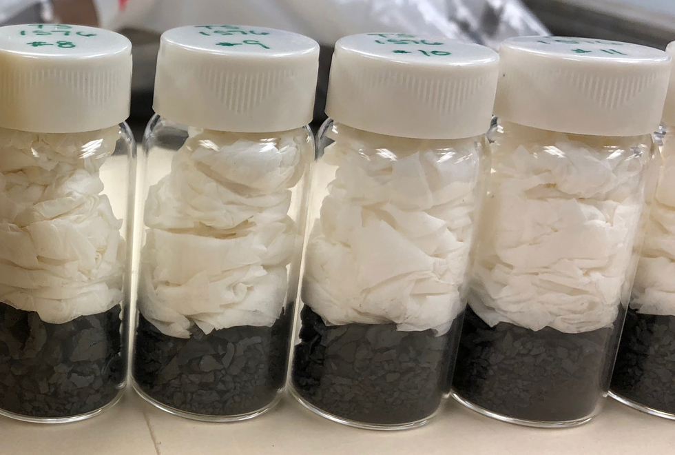 A row of clear vials with white plastic caps on a countertop. Each vial contains a pile of moist wipes on top of small metal chunks.