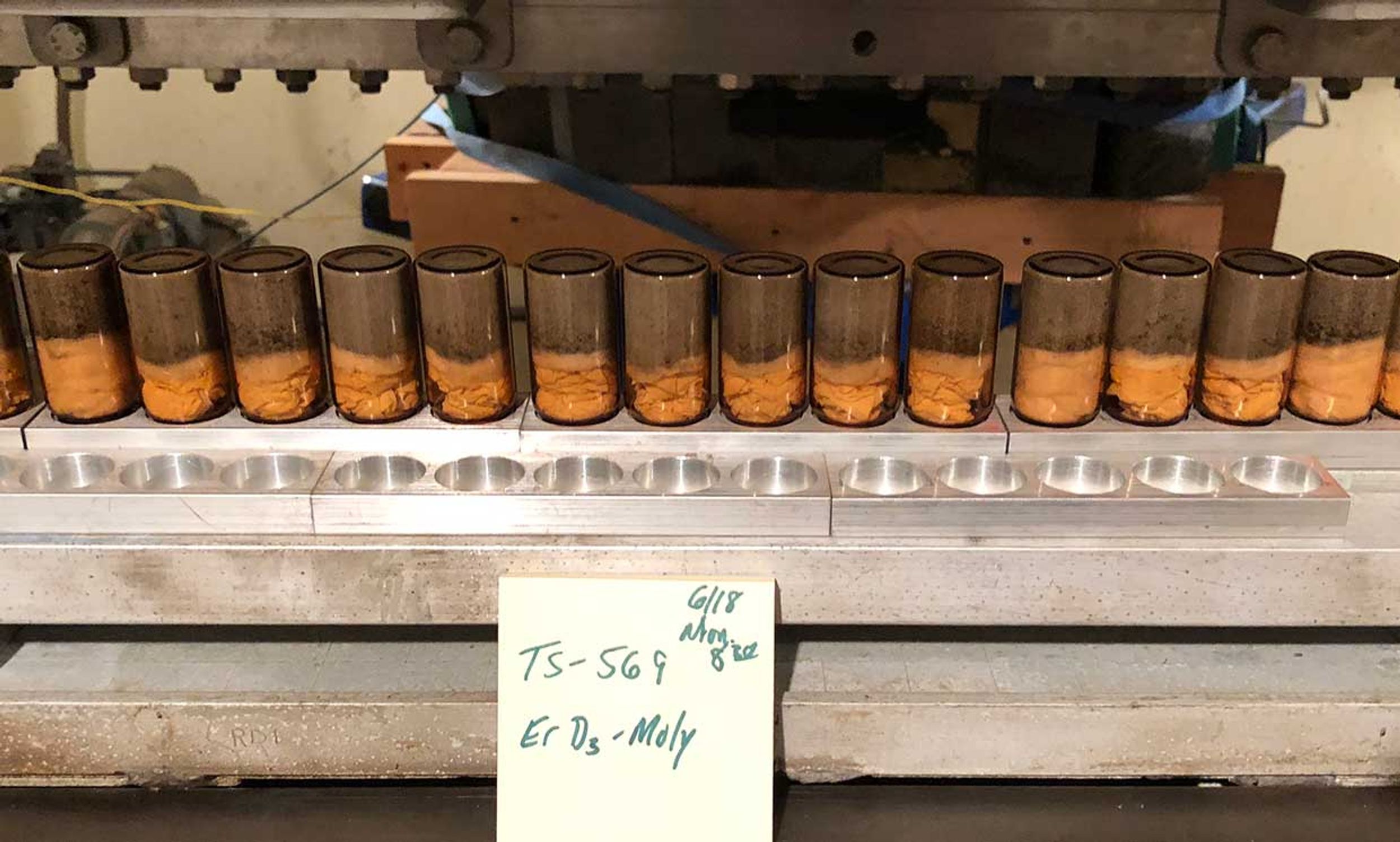 A row of orange tinted powdery samples sitting in the bottom halves of a row of transparent cylinders