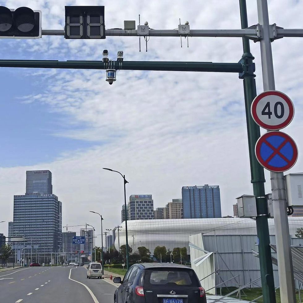 A scene from a multilane road has buildings in the background and the back of a car in the foreground. Along the rightmost lane there are two roadside structures, a white one bearing standard signage and a green oneu2014consisting of a vertical pole and a horizontal cross beam at the top of the pole. On the white pole can be seen a box; another such box is on the vertical green pole; and on the topmost green crossbar there are sensors.