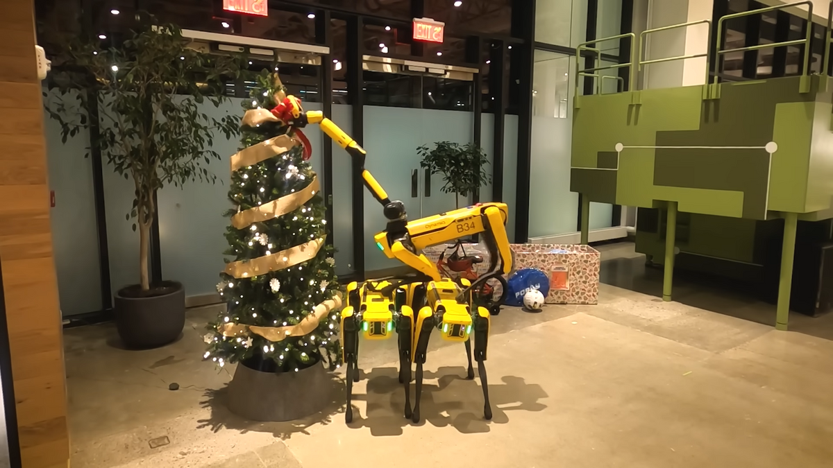 A screenshot of a video showing a pyramid of three yellow quadruped robots putting a bow at the top of a Christmas tree
