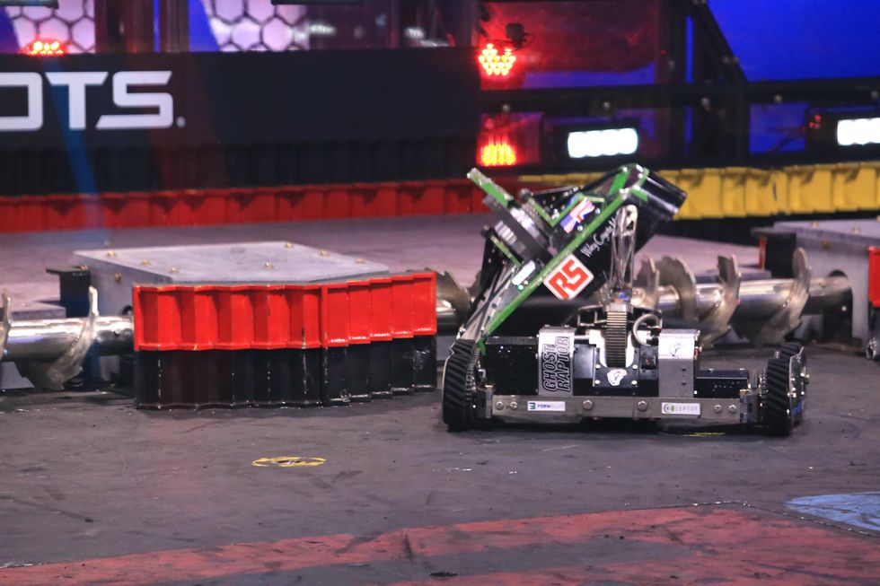 A shiny wheeled  and low-slug robot pushes a black and green low slung robot into a horizontal shredding device in an arena