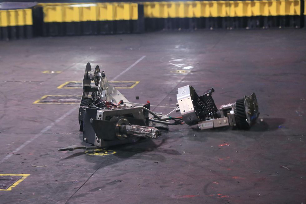 A shiny wheeled robot lies in large fragments, connected by some electrical cables, on an arena floor
