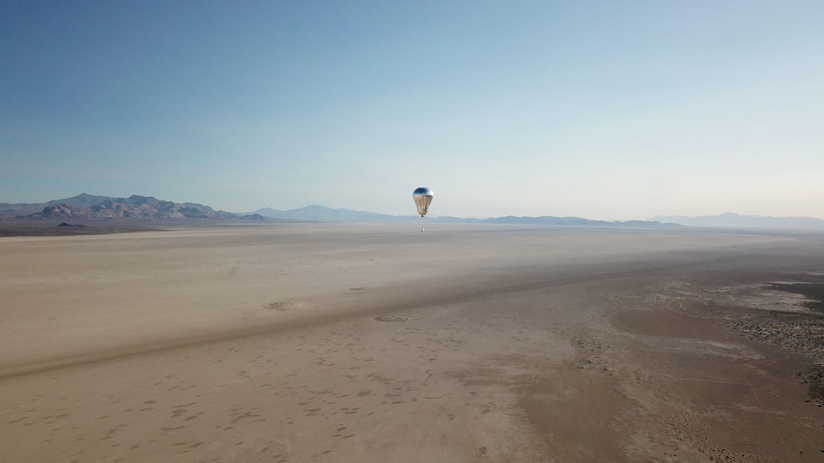 A silver balloon with a robotic payload drifts high above a desert landscape