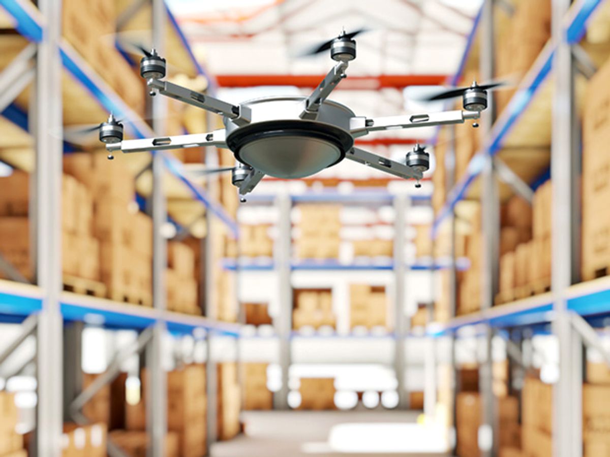 A silver drone flies through an aisle of sheleves lined with boxes in a warehouse.
