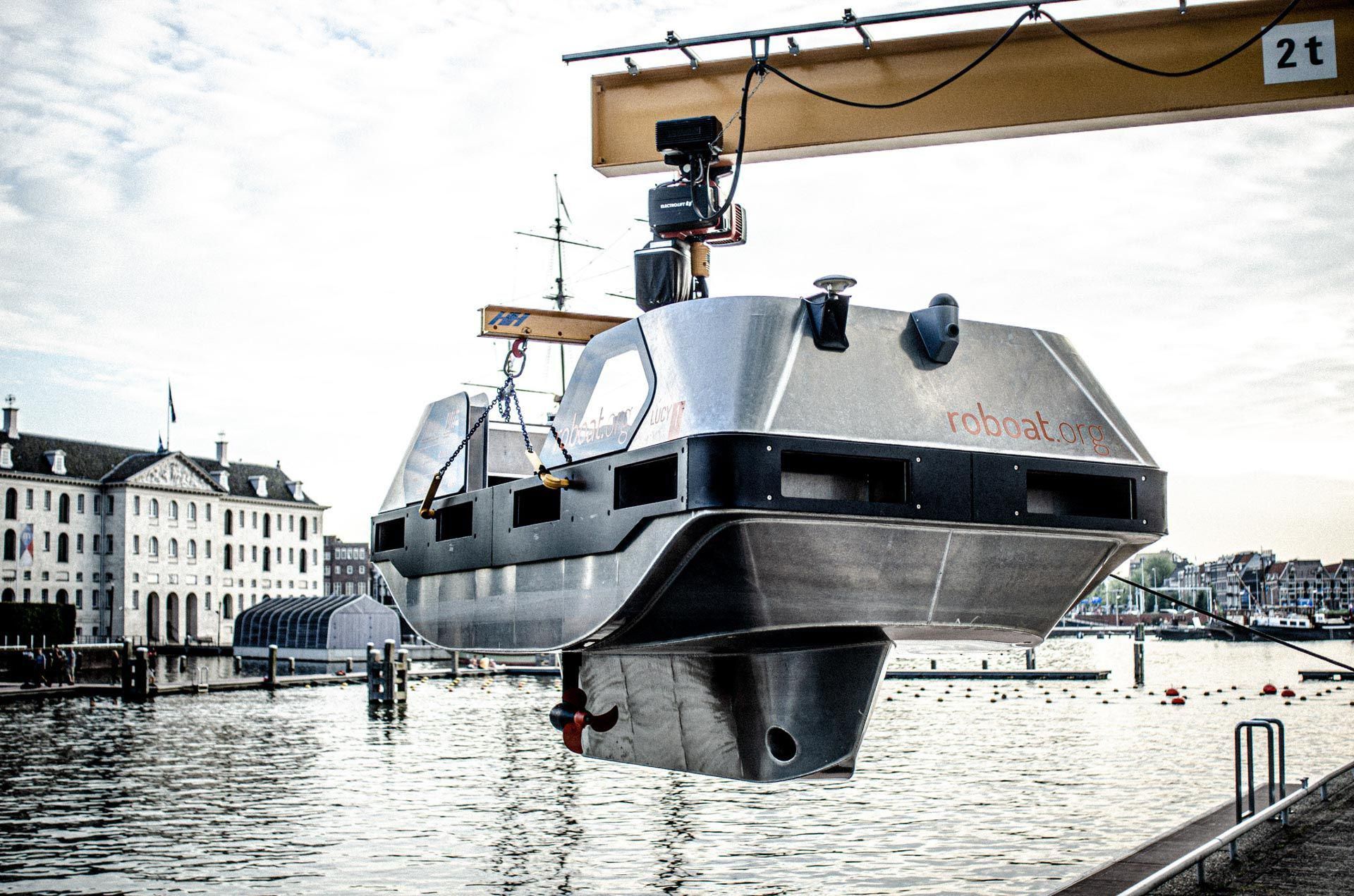 A silver robotic boat hangs from a yellow crane over an Amsterdam canal