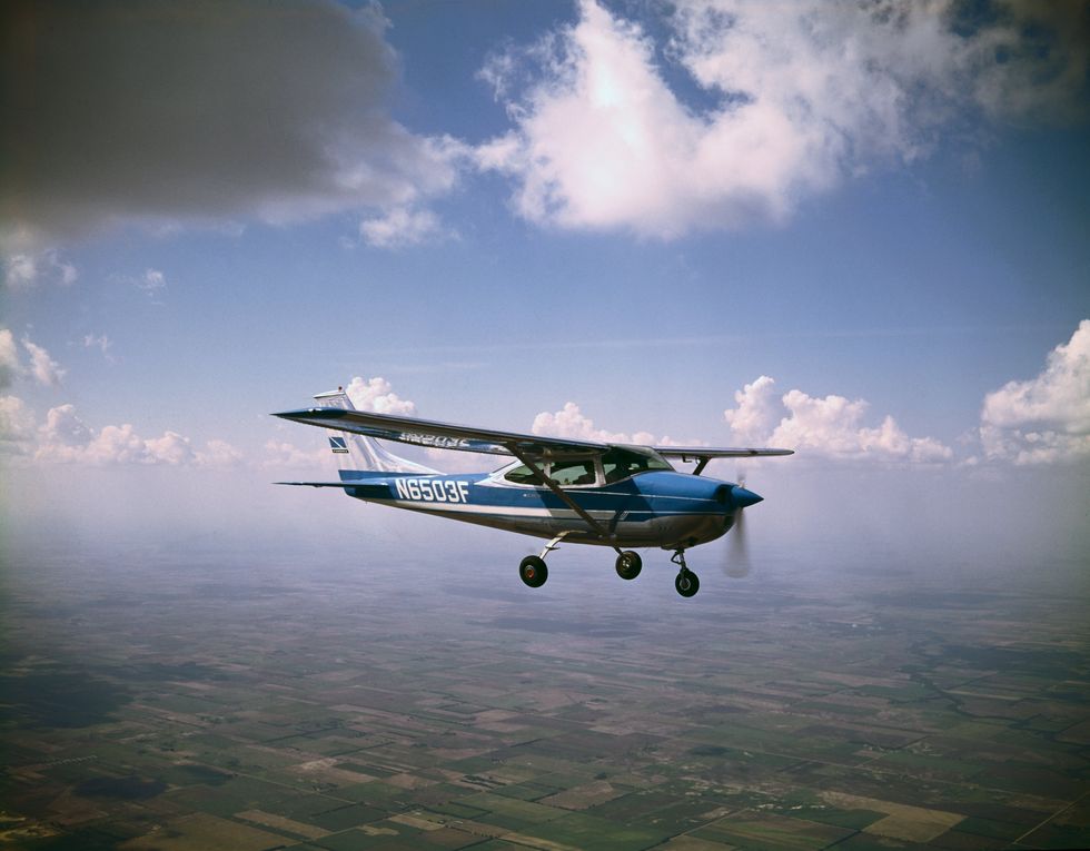 A small, blue-and-white airplane with a single propeller flies under clouds and over farmland