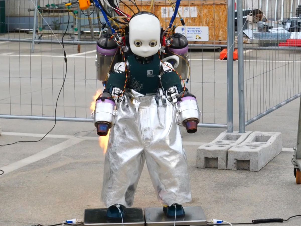 A small humanoid bipedal robot with a jetpack, jet arms, silvery flame-protective pants and many wires stands on two bathroom scales.