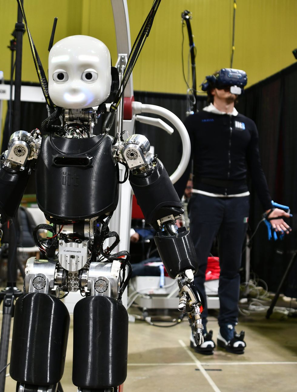 A small humanoid robot stands in the foreground while a human in a VR headset stands in the background, controlling the robot through his motions.