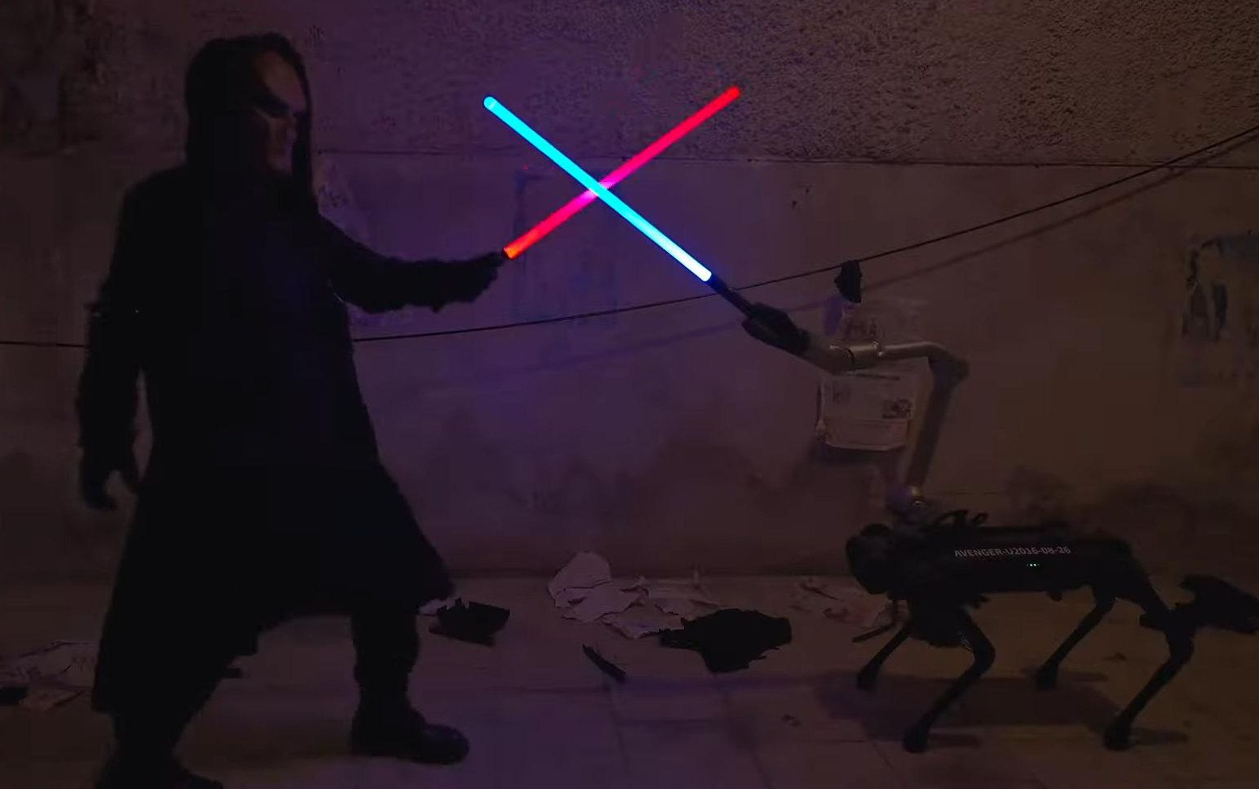 A small quadrupedal robot with an arm on its back holds a blue light saber against the red light saber of a human in a mask dressed in black
