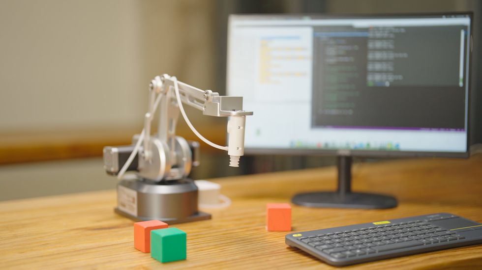 A small silver metal robotic arm the size of a desk lamp stands on a wooden desk next to small red and green wooden blocks, a computer keyboard, and a monitor.