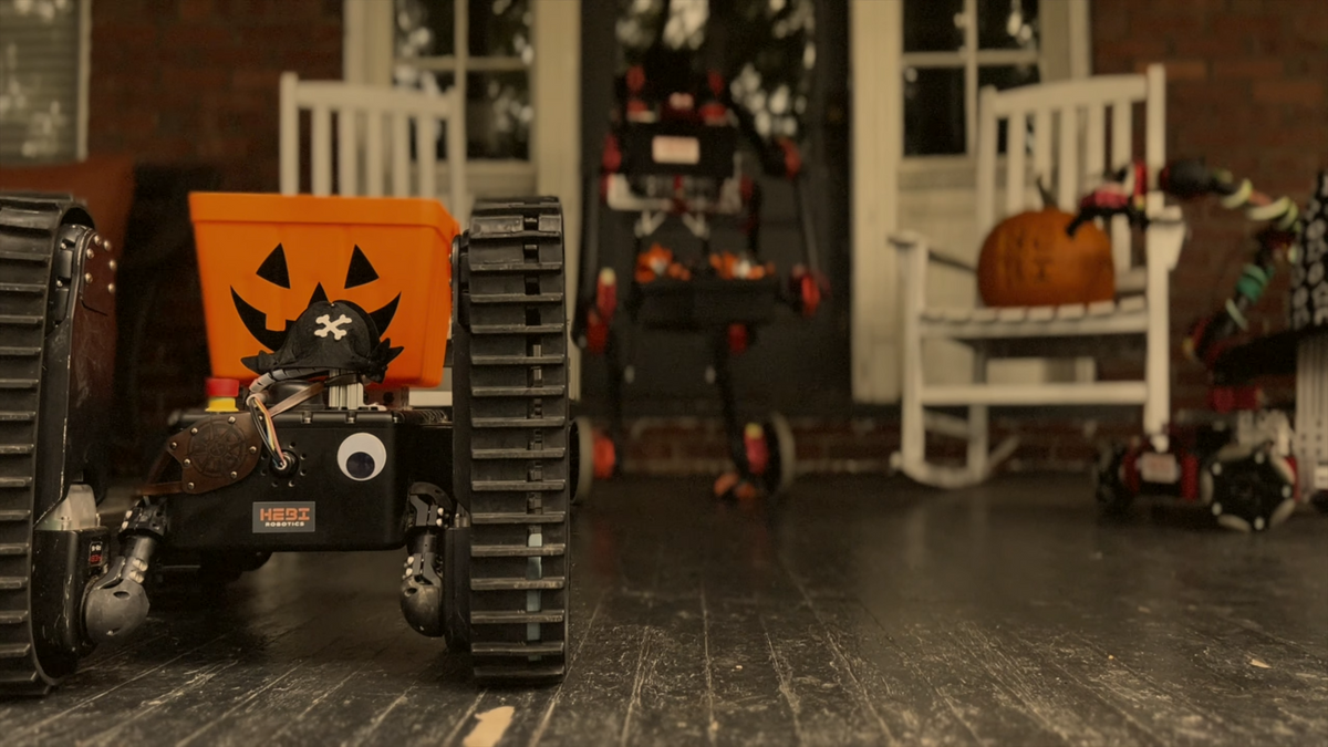 A small tracked robot in a pirate costume with a pumpkin bucket on its back on a Halloween decorated porch with two other robots out of focus in the background