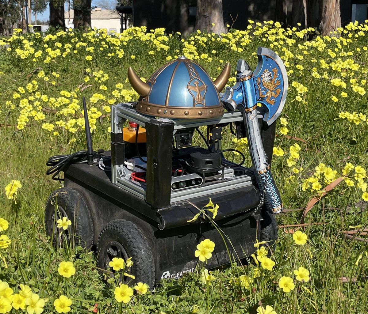 A small wheeled robot in a field of yellow flowers with a toy Viking helmet and axe placed on it