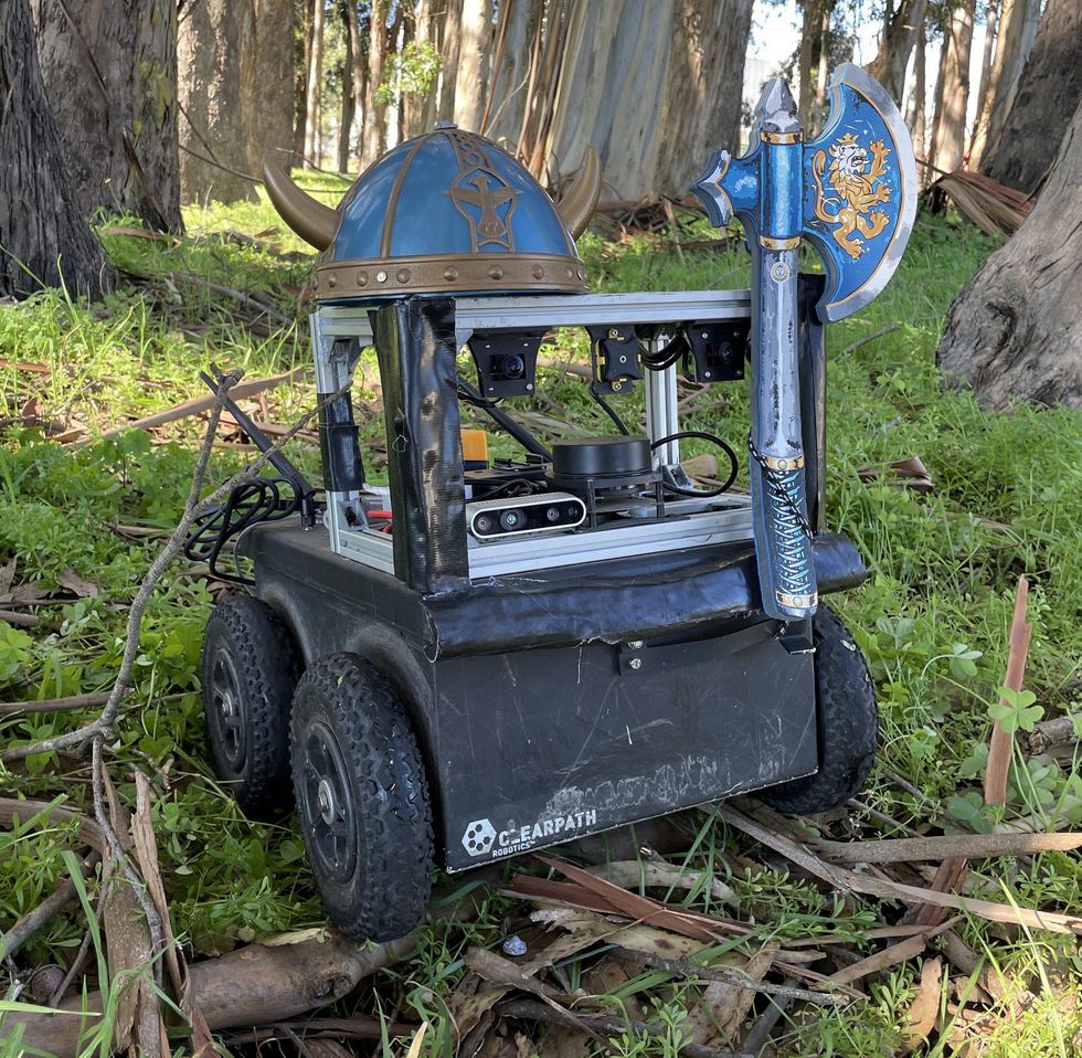A small wheeled robot in a forest with a toy Viking helmet and axe placed on it