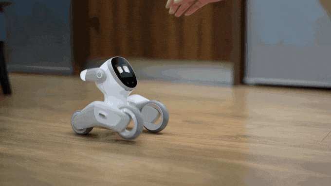 A small white robot with four wheels, moving ears, and an expressive animated face reacts cutely to being pet by a human