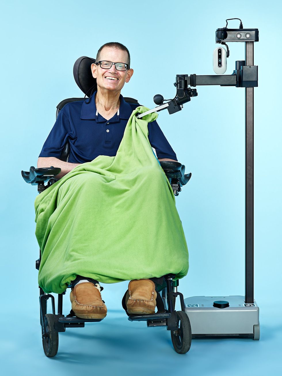 A smiling bespectacled man in a wheelchair is seated next to a robot consisting of a mobile base, a thin vertical pole, and a horizontal arm, whose gripper is repositioning a green blanket on the man\u2019s lap.