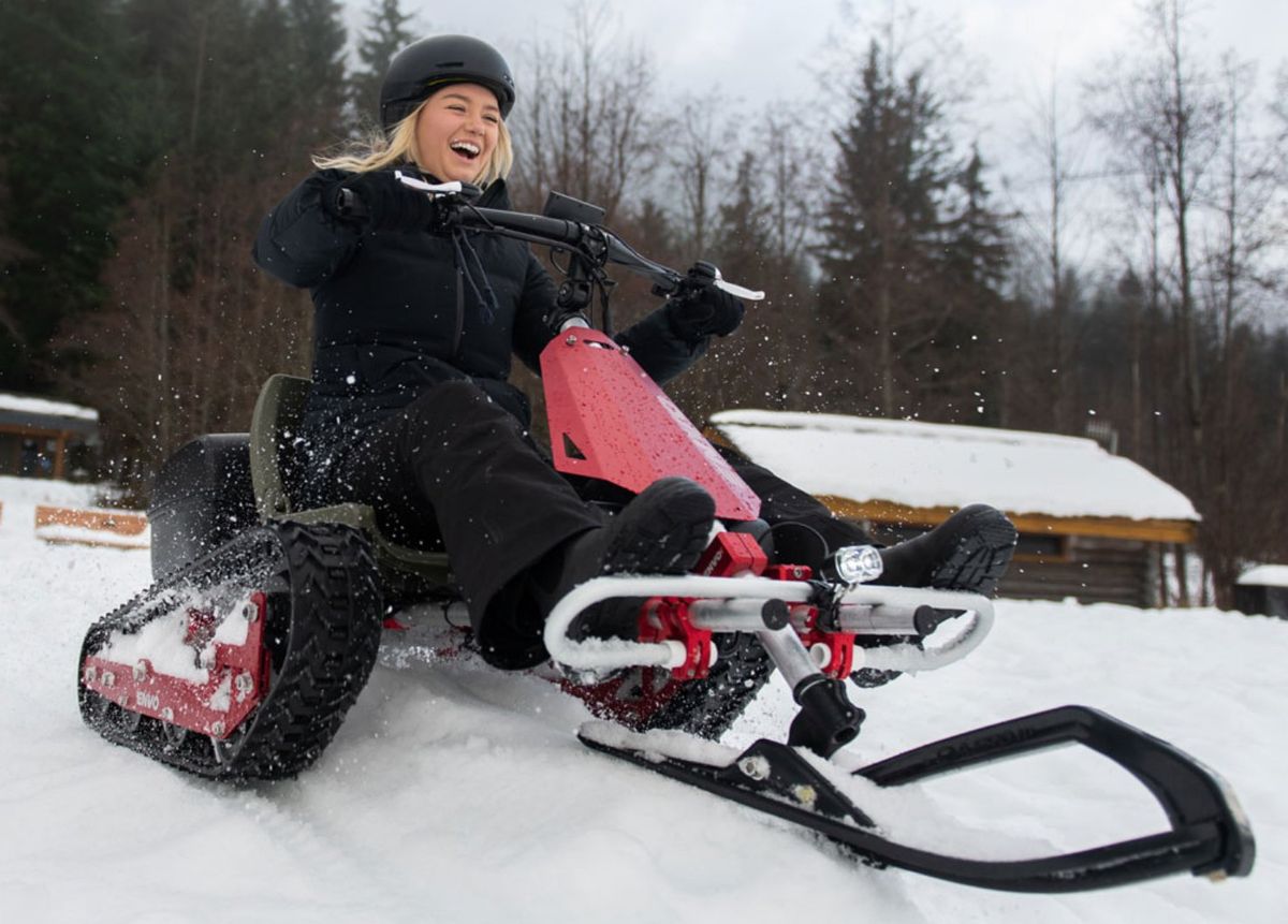 A smiling blonde woman in a helmet and snow gear rides a black and red snowmobile.