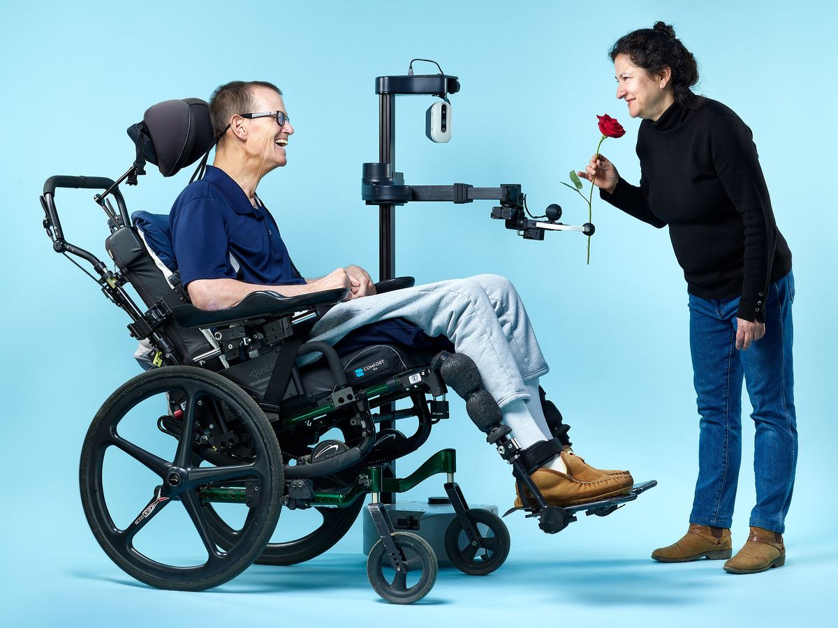 A smiling man in a wheelchair connects eyes with a smiling woman in jeans and a turtleneck, who bends over to smell a rose that is in the gripper of a tall, pole-thin, simple robot. 