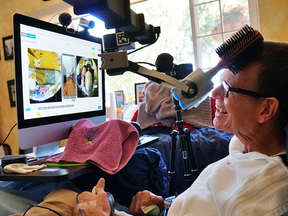 A smiling man lies in bed. He is looking at a monitor which shows multiple camera views, including one of himself, as a robotic gripper holding a hairbrush scratches his head.