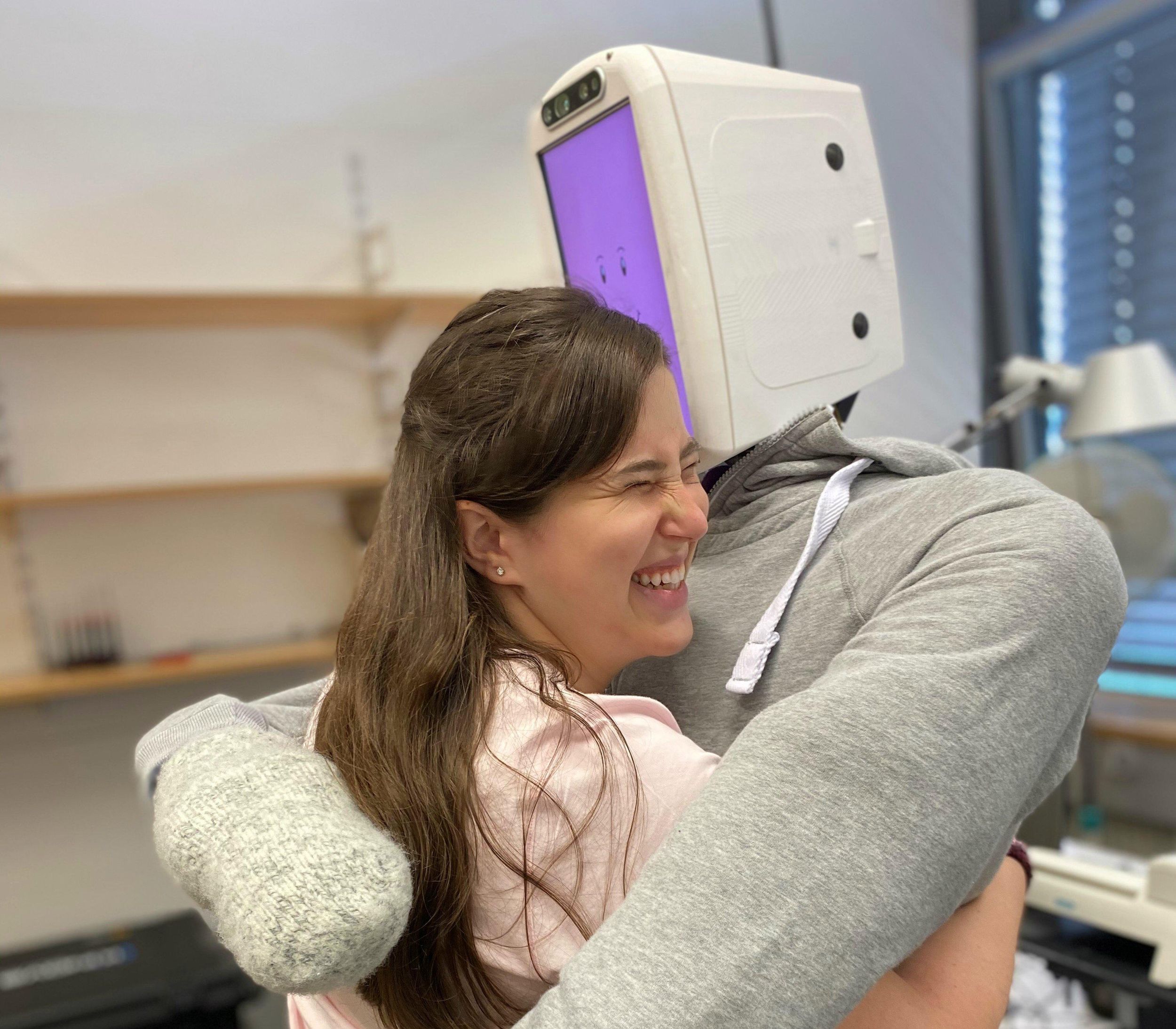 A smiling woman hugs a robotic torso with a soft grey sweatshirt. The robot's head has a purple computer screen with a smiling face.