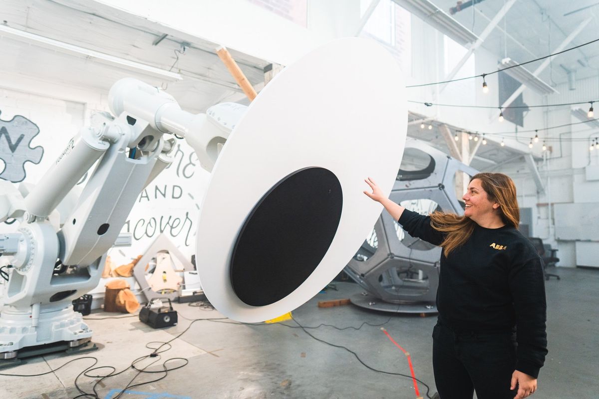 A smiling woman stands next to a robot arm with an enormous googly eye mounted on it