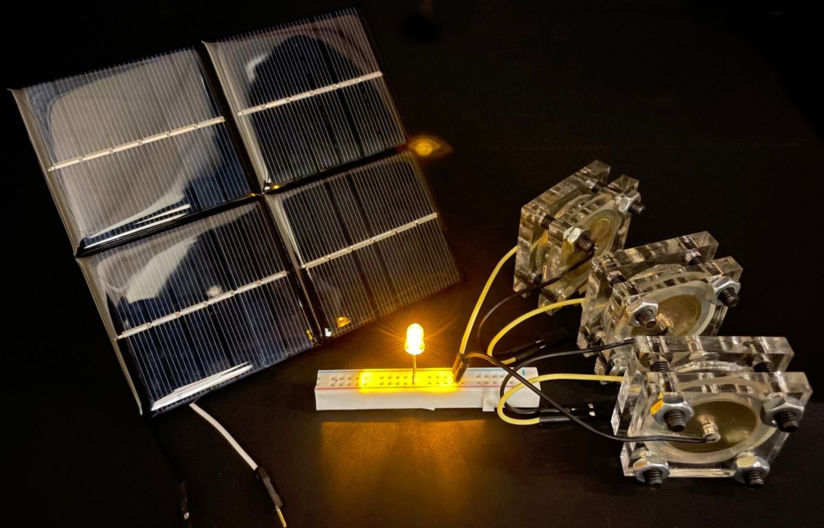 A solar panel sits on a table next to a set of three square devices plugged into a small bread board with a glowing yellow LED light.