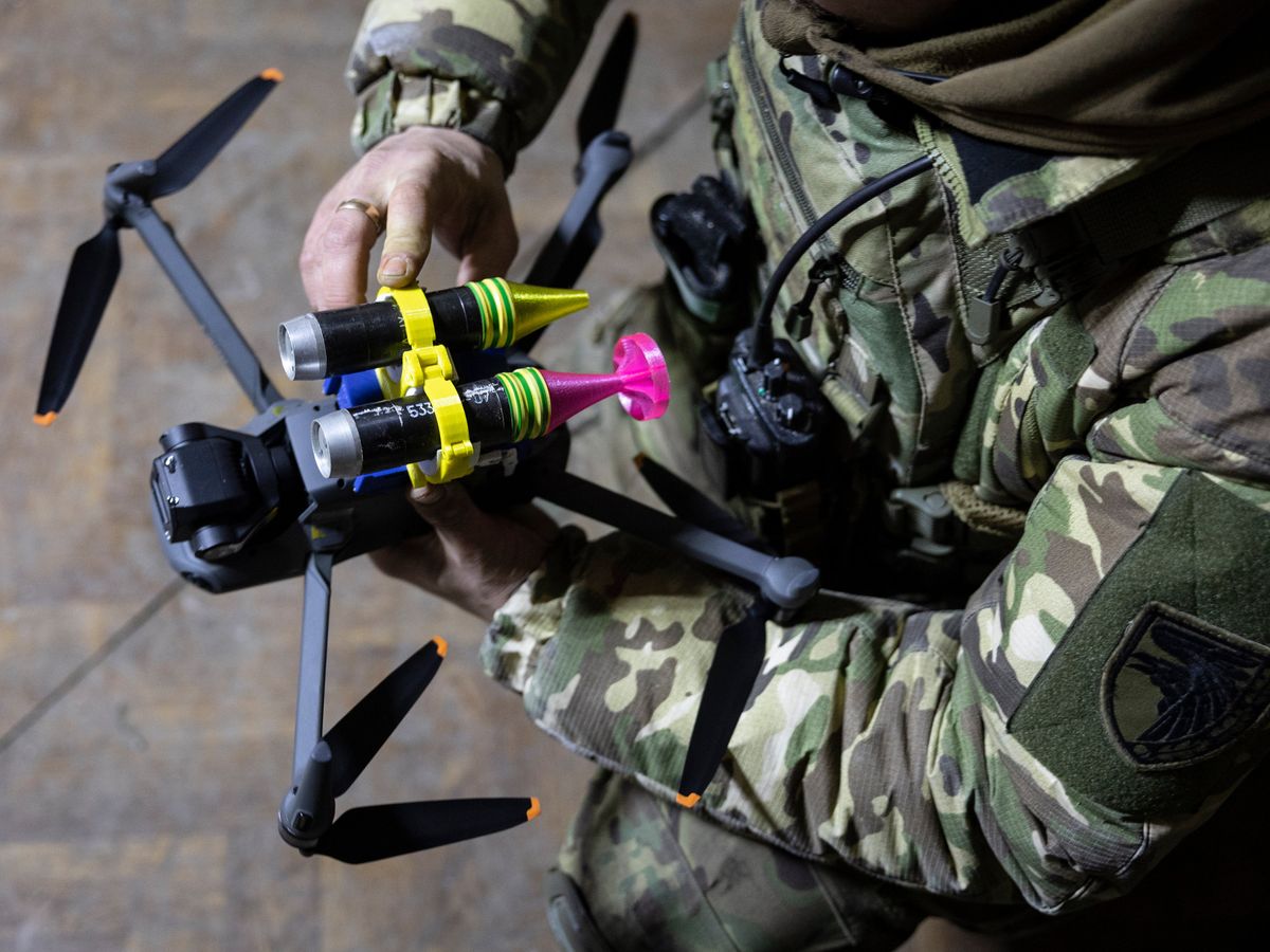 A soldier in fatigues holds a black quadcopter drone, which has been equipped with two grenades attached with colorful 3D printed holders.