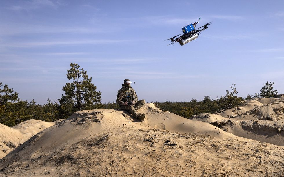 A soldier sits on a sandy hill wearing special glasses and holding a remote to control a drone with a fake bomb which is in the air in front of him.