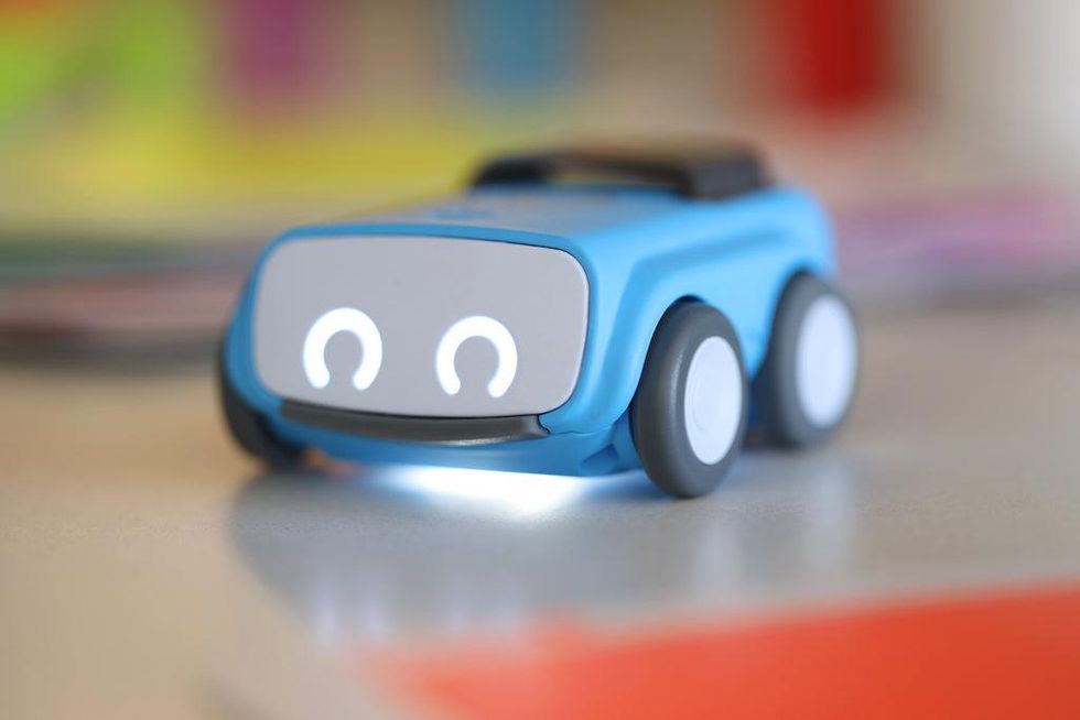 A Sphero Indi robot, which has a rectangular blue body with four wheels and two circles as eyes, stands on a colorful desk.