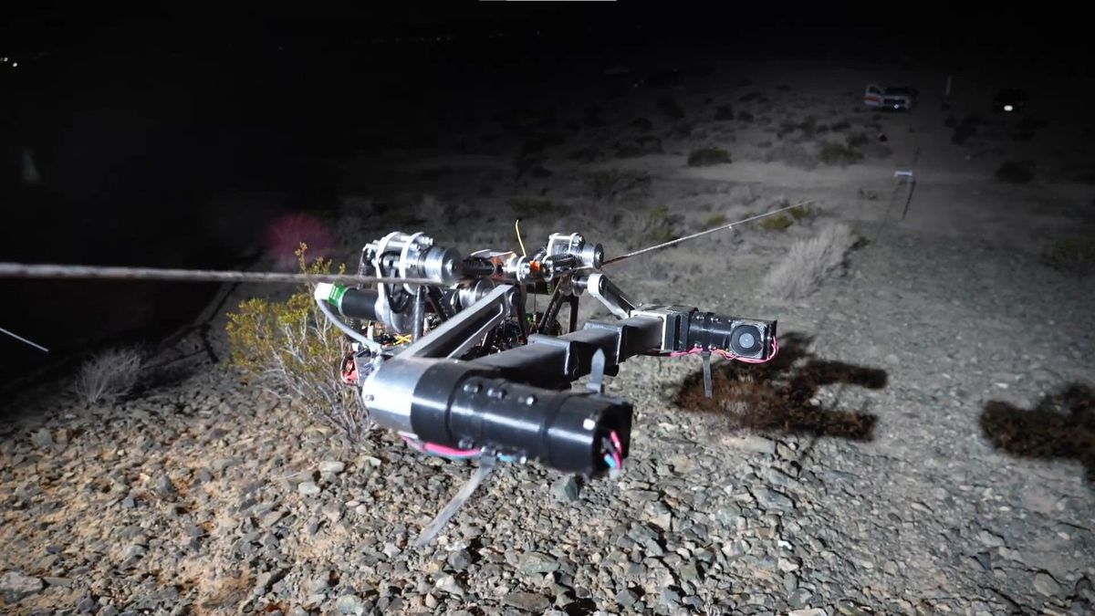 A spindly robot made of cables and actuators slowly travels down a cable above a desert landscape.