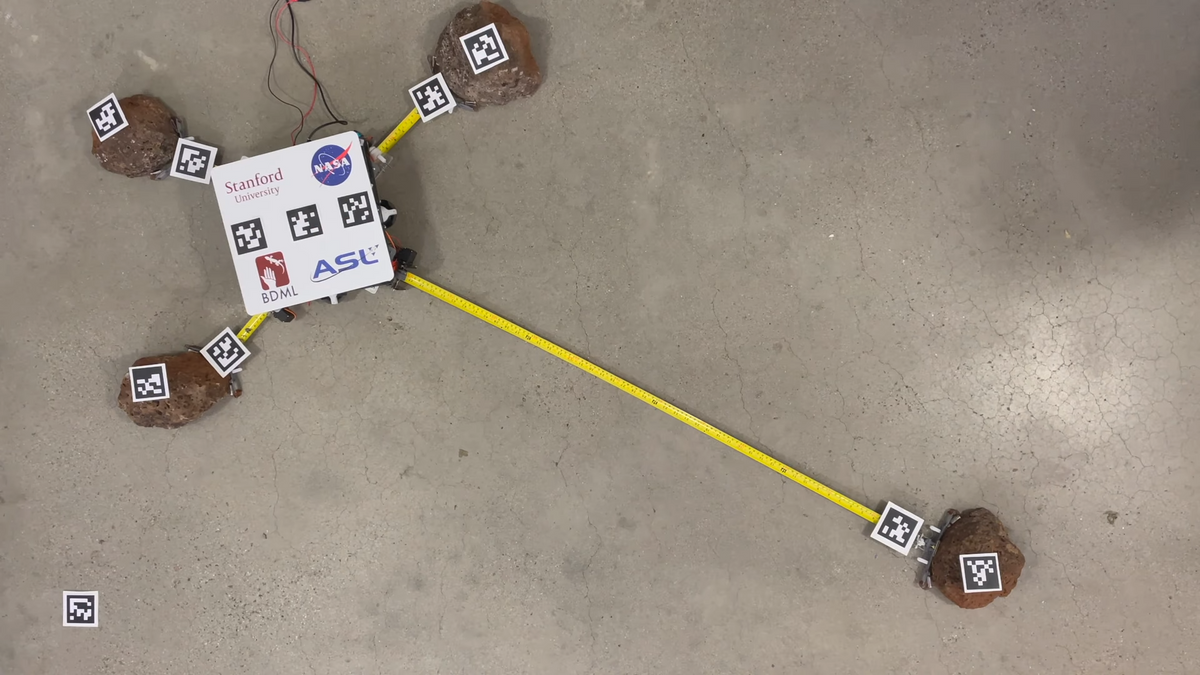 A square white robot on a concrete floor attaches to rocks using grippers on the ends of extended metal measuring tapes.