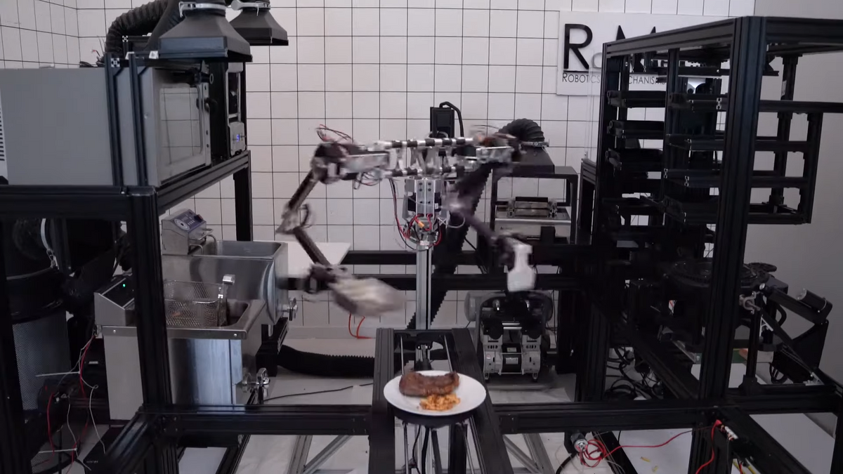 A still from a video shows a metal robot with arms surrounded by kitchen equipment. 