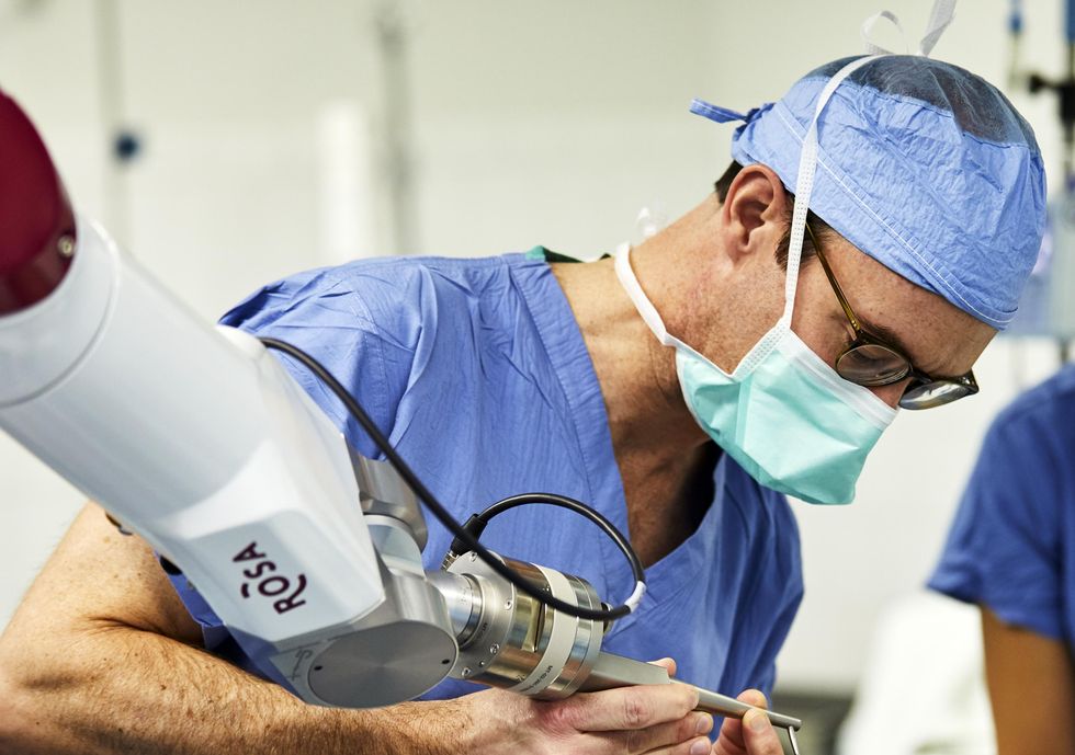 A surgeon wearing scrubs and a facemask holds the end of a robotic surgical tool.