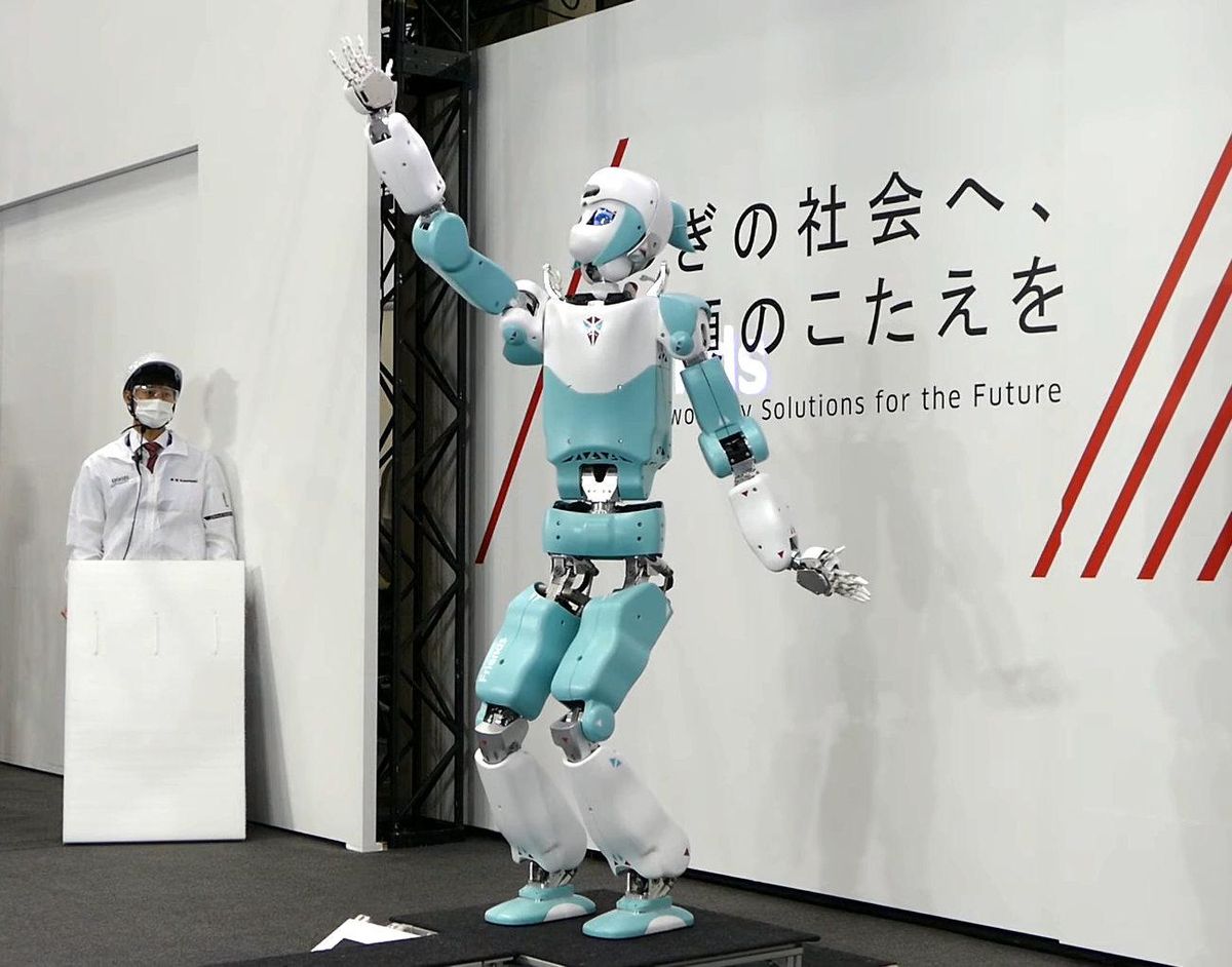 A teal and white humanoid robot stands on a stage with its arm outstretched