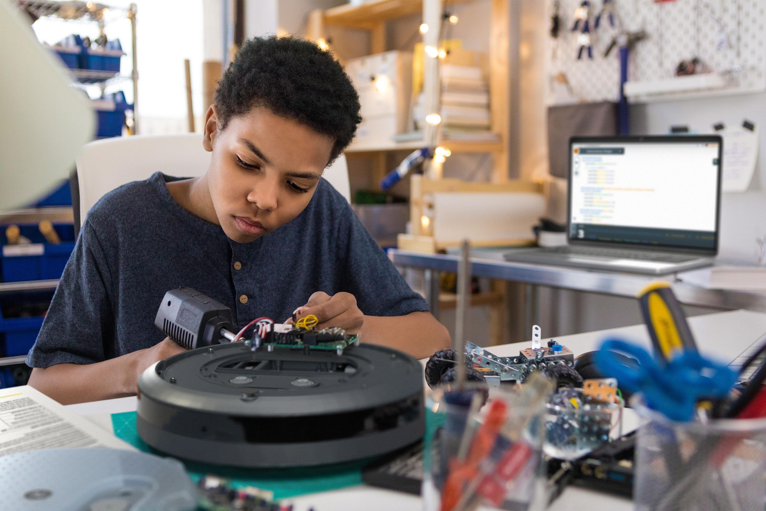 A teen adds electronics to a Roomba on a cluttered workbench