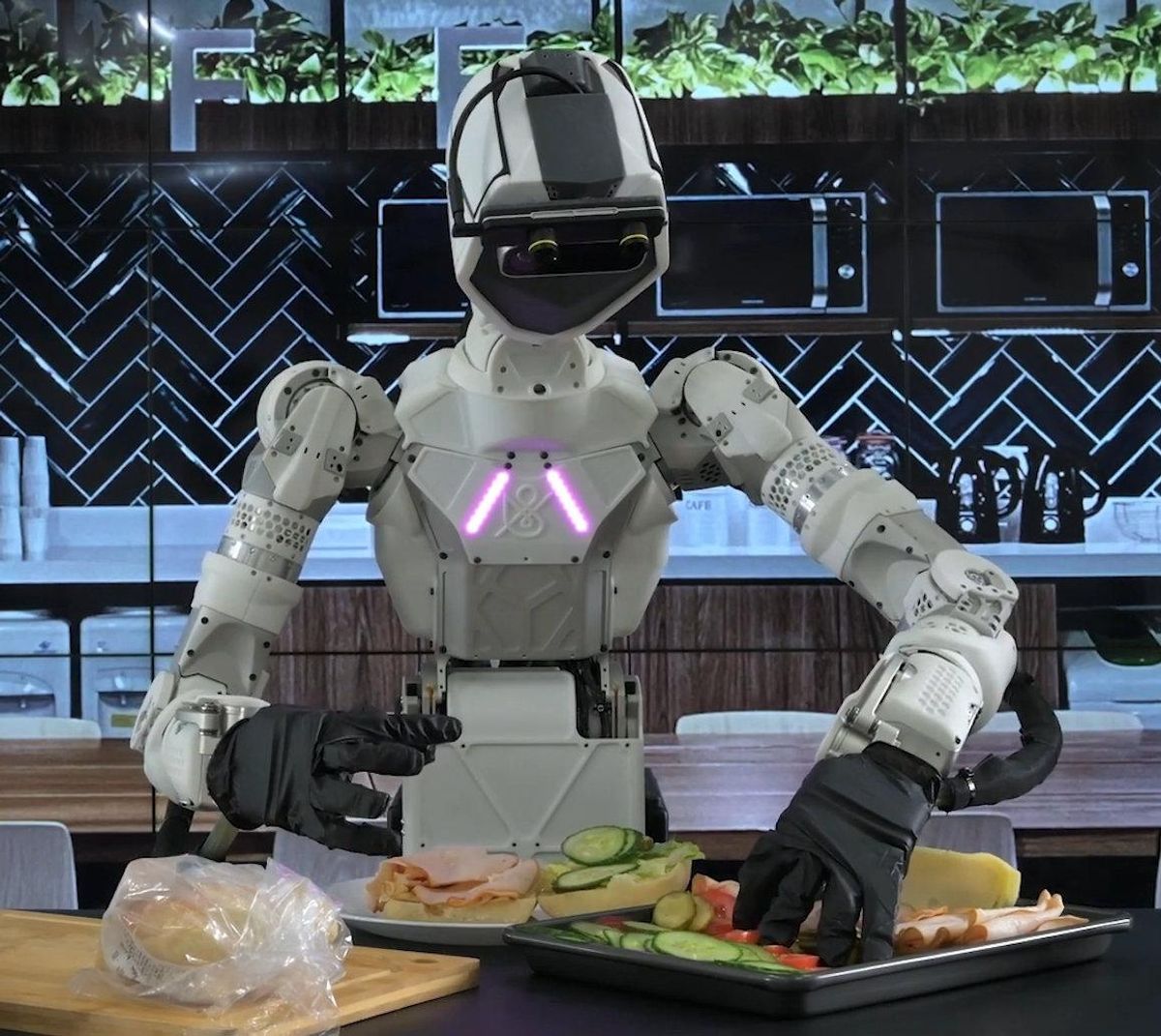 A teleoperated humanoid robot torso stands in a kitchen assembling a turkey sandwich from ingredients on a tray