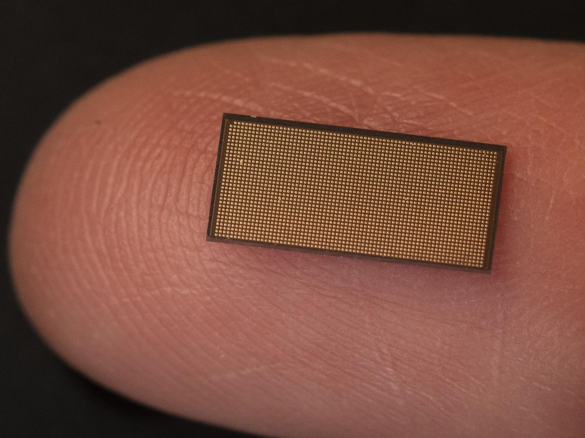 A tiny chip rests on a fingertip. It is rectangular with a dense matrix of tiny gold dots.