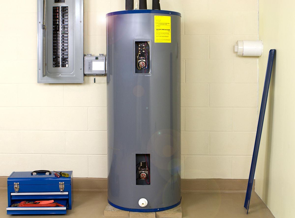 A water heater in a basement with a fusebox and blue tool box.