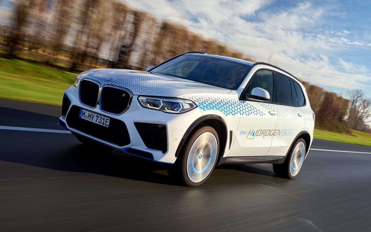 A white car that says BMW Hydrogen on the side drives through a landscape with grass and trees.