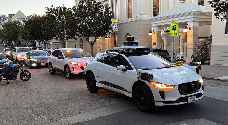 A white car with running lights on and with the word \u201cWaymo\u201d emblazoned on the rear door stands in a street, with other cars backed up behind it.