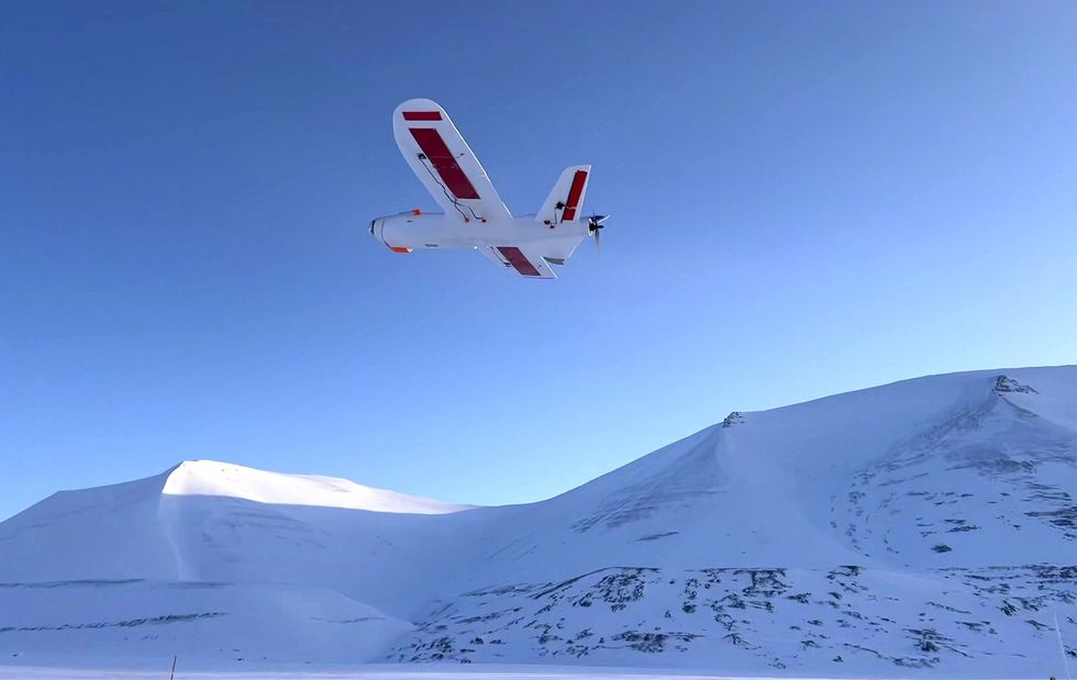 A white drone with red markings flies above a snowcovered hill against a blue sky.