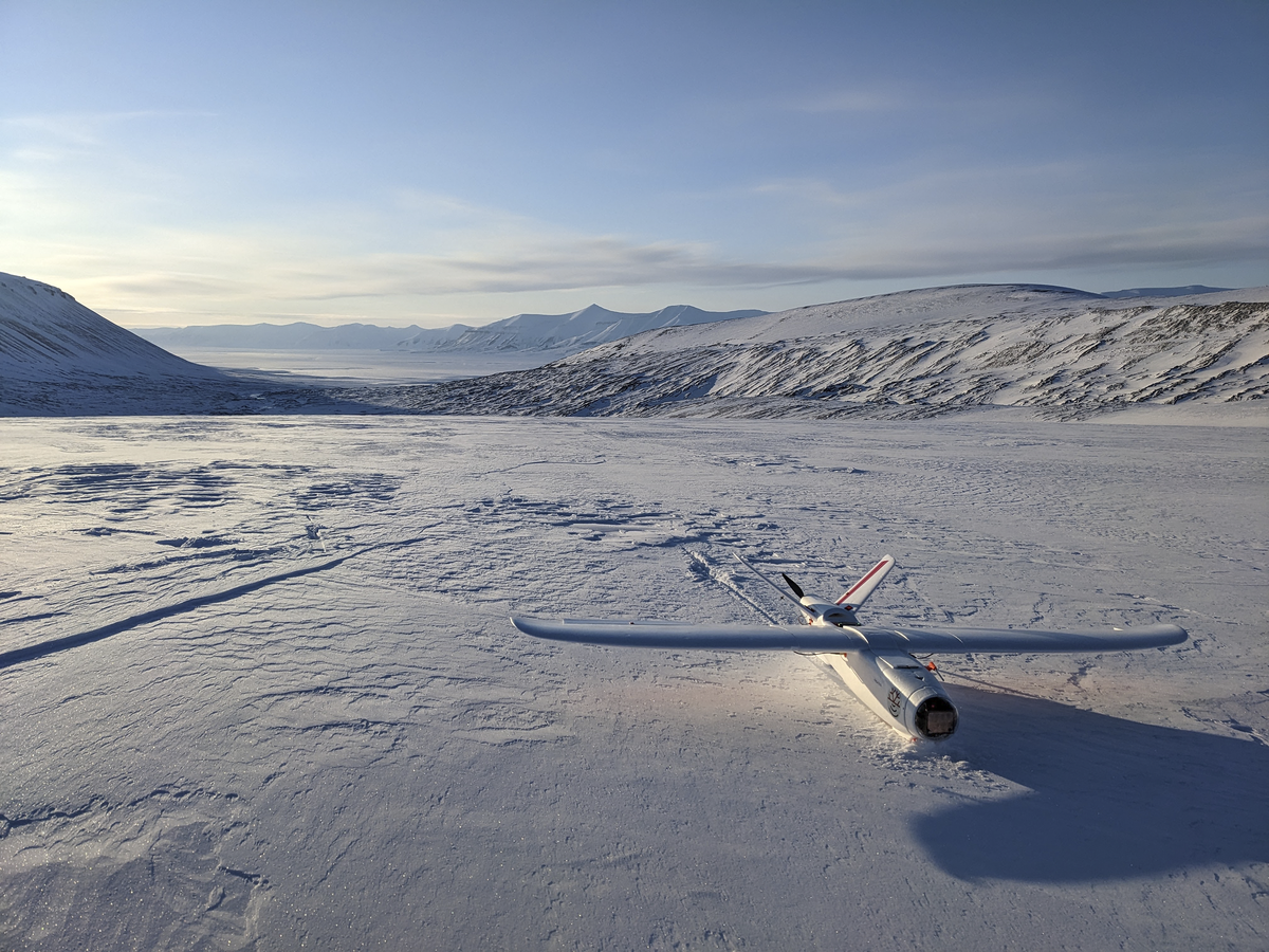A white drone with red markings lands on a plain of snow and ice, snow-covered hills in the background.