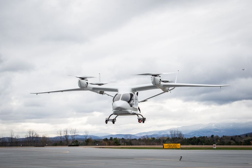A white eVTOL aircraft is shown hovering about 10 meters above an airport apron during a cloudy day