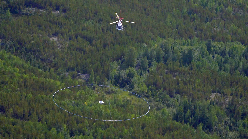 A white helicopter carrying a large loop many times its size beneath it flies over an evergreen forest.