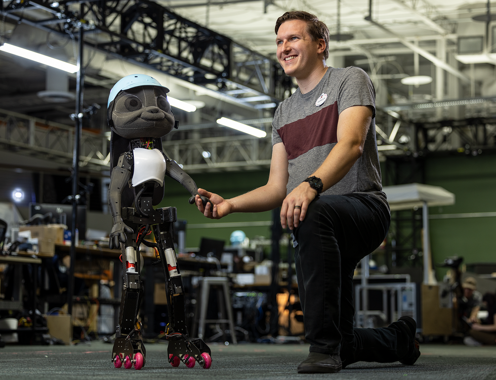 A white man bending down to shake the hand of a child-like robot wearing a white hat and white vest.