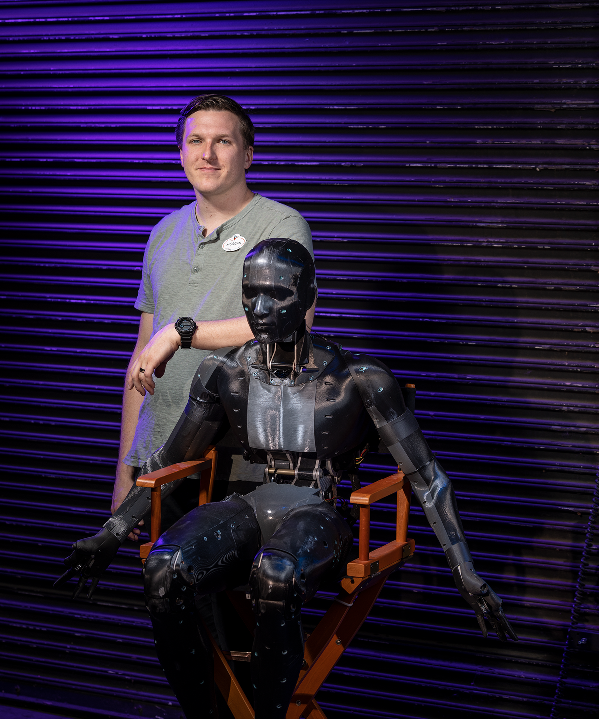  A white man standing alongside a human-looking robot seated in a red chair. 