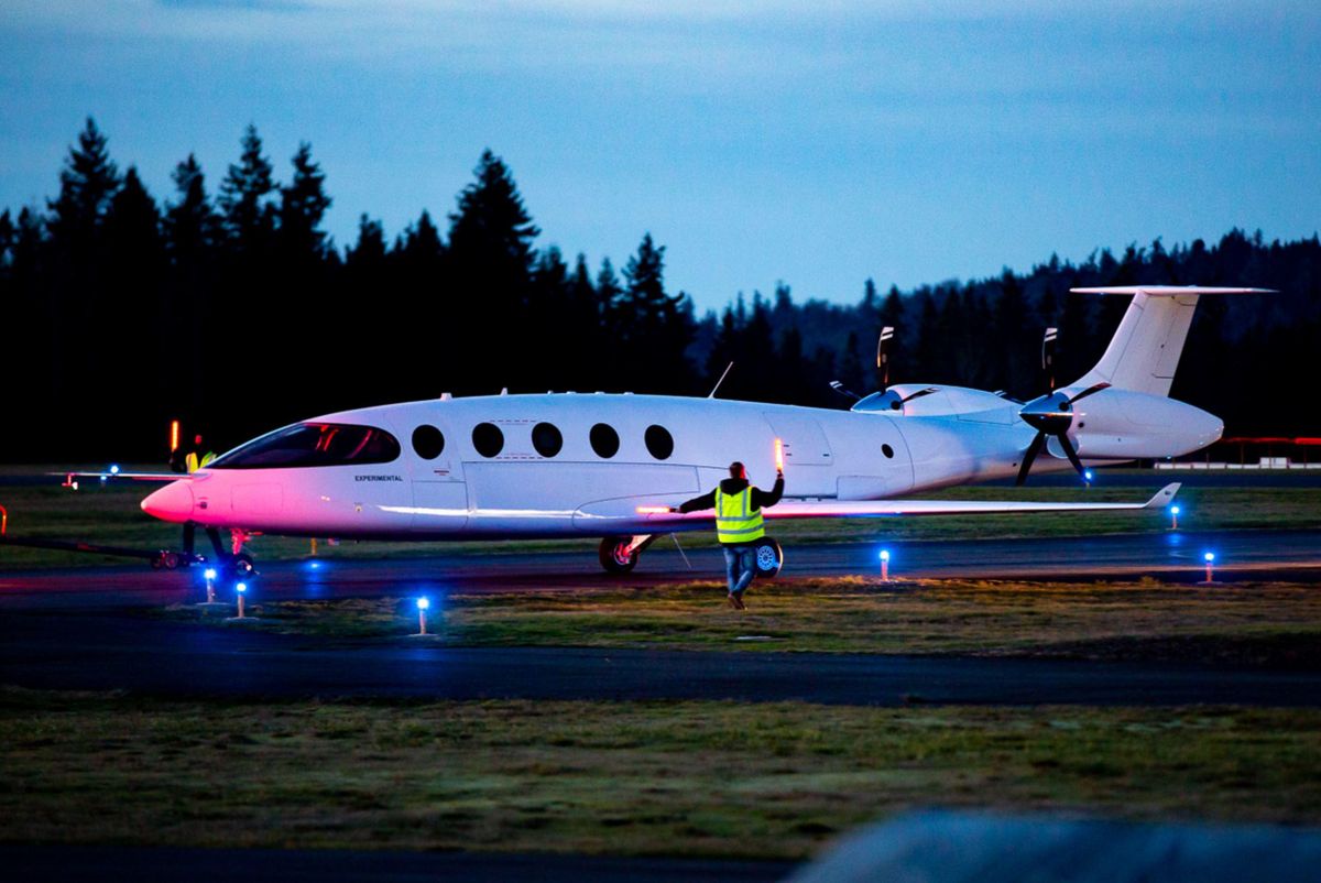 A white nine-seater aircraft sits on a runway at dusk. An airport marshaller in a reflective vest holds up two light sticks in front of it.