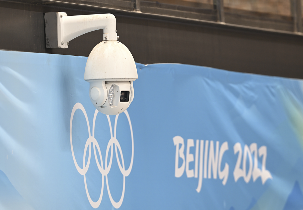 A white ovoid hangs from a horizontal fixture attached to a wall, in front of a blue flag bearing the five-ring Olympic logo, next to the label, u201cBeijing 2022u201d.