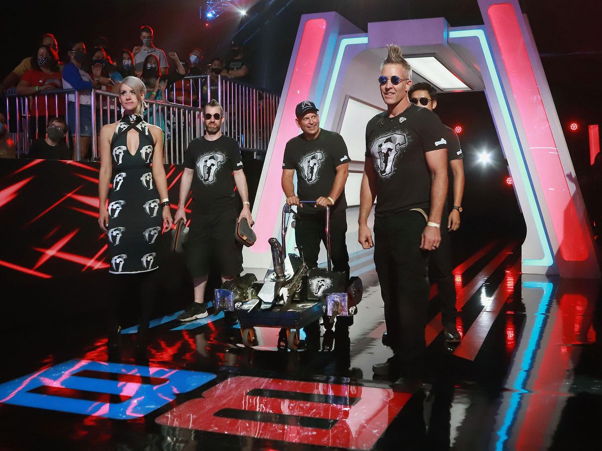 A woman and four men surround a gleaming tracked robot on a push cart. They are standing in a brightly lit entrance to an  arena. The woman is wearing a black bodycon dress and the men are wearing black t-shirts all with a stylized animal skull logo