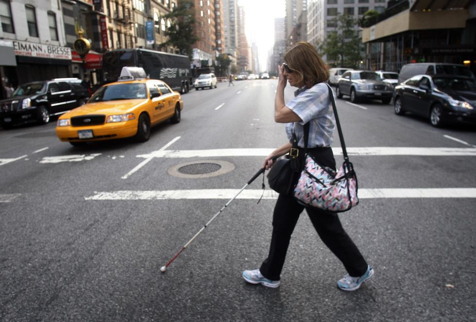 A woman crosses a street in New York City using a white cane for assistance.