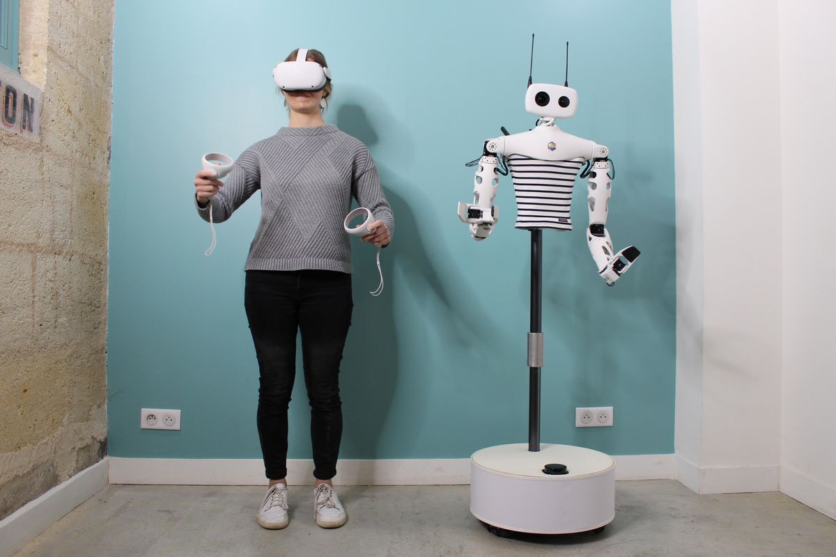A woman in a VR headset holding motion controllers stands next to a humanoid robotic torso on a mobile base